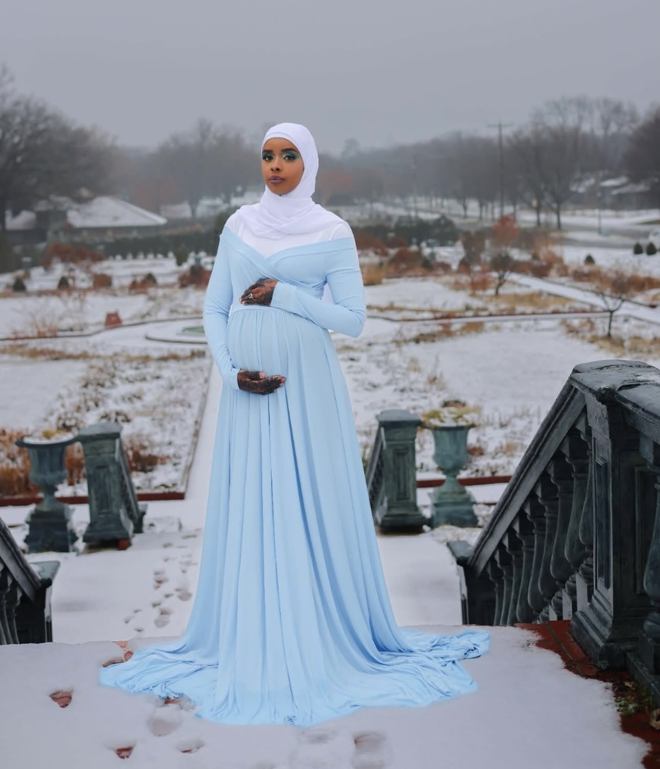 Expecting mother in a flowing powder-blue gown and white hijab standing on snow-covered garden steps